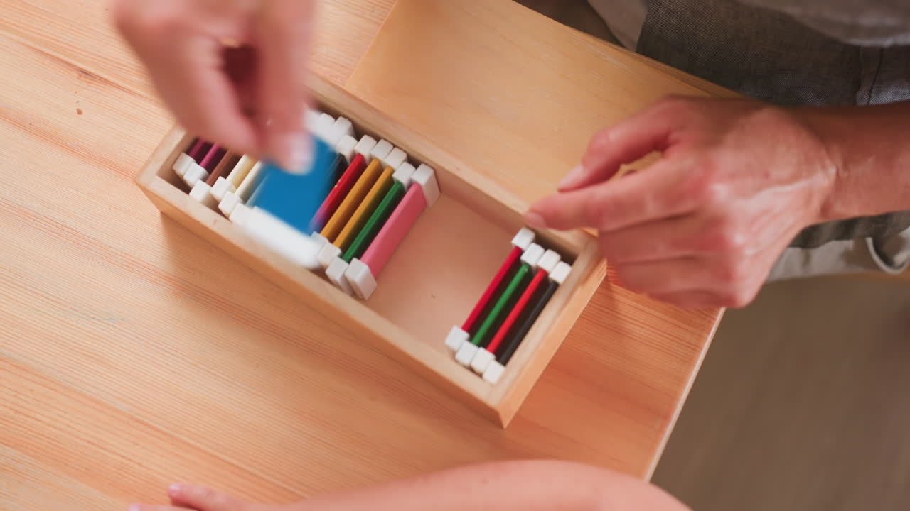 Adult arranges colorful blocks back into wooden box while kid assists beside, finger pointing at organized pieces, supportive educational activity in classroom setting promoting order