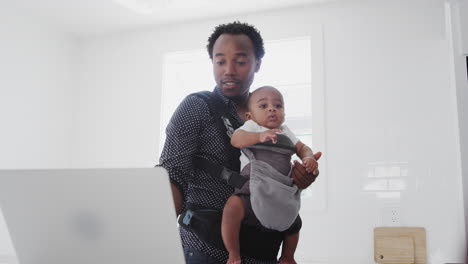 father with baby son in sling multi-tasking working from home on laptop