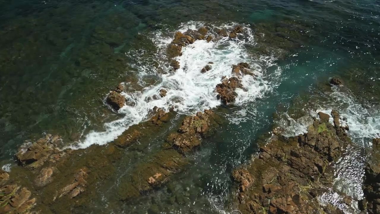 Una ripresa con drone di onde blu che si infrangono contro rocce marrone-rossastre sulla costa mediterranea in Francia. Il sole luccica sull'acqua mentre le onde si infrangono contro le rocce.