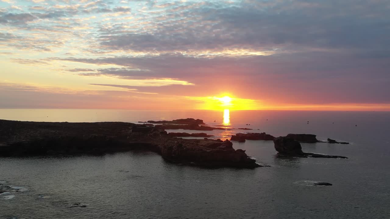 Plano cinematográfico com drone com vista para silhuetas de penhascos rochosos e o Oceano Atlântico durante um pôr do sol vibrante, com a luz do sol refletindo na superfície da água