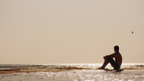 Einsame-Silhouette-Eines-Jungen-Mannes,-Der-Auf-Dem-Sand-Am-Meer-Sitzt