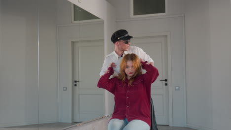 man in uniform holds up hands of seated woman in red shirt as he dances playfully behind her, both reflected in large wall mirror inside bright modern room with white doors