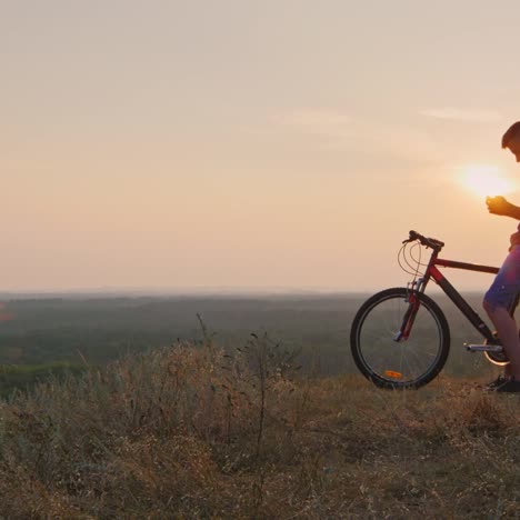 A-young-man-uses-a-smartphone-stands-by-his-bike-at-sunset