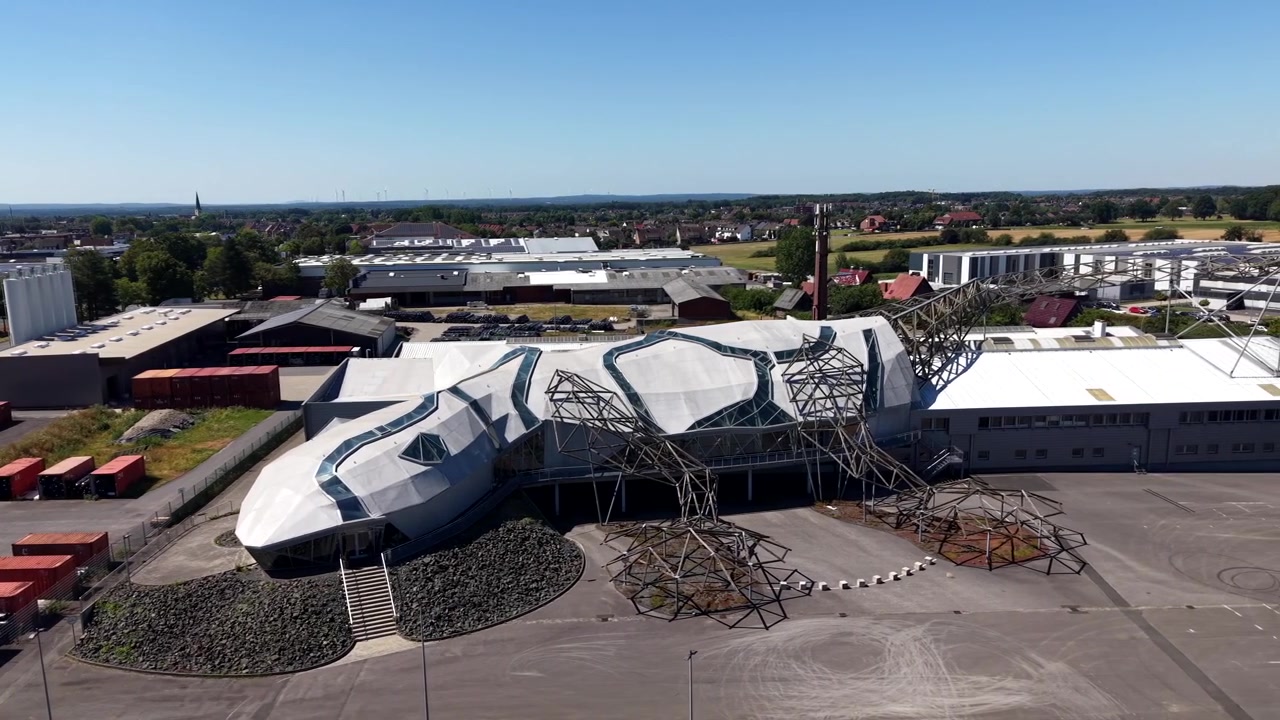 Eckiges, geckoartiges Gebäude mit facettierten weißen Dachpaneelen neben einer skelettartigen Stahlfachwerkkonstruktion in Dülmen, Deutschland; gestapelte Schiffscontainer auf Industriegelände. Seitliche Weitwinkel-Luftaufnahme mit Drohne