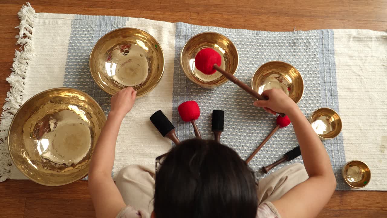 Top-down view of hands gently playing a set of brass Tibetan singing bowls arranged on a woven mat in a calm indoor wellness studio — ideal for meditation and sound healing