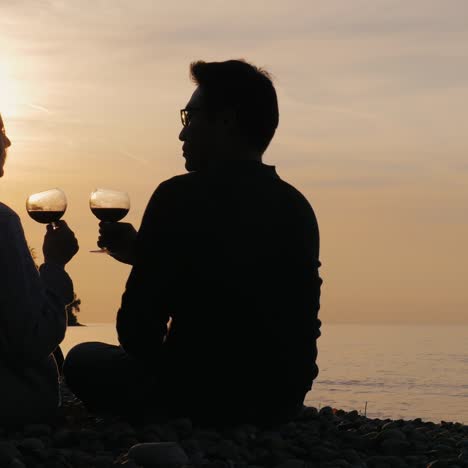 Silhouetten-Eines-Jungen-Paares,-Das-Wein-Bei-Sonnenuntergang-Am-Ufer-Eines-Sees-Trinkt