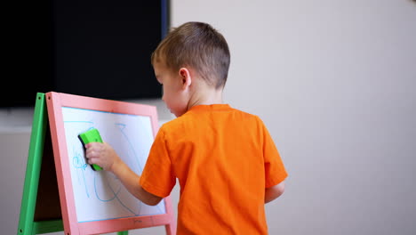 baby boy in orange t-shirt has drawn a picture on whiteboard. kid takes a duster and cleans the easel.