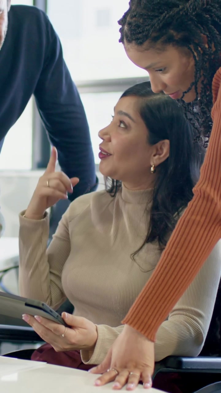 Vídeo vertical: Mulher sentada sorrindo e tocando tablet na mesa, com blusa bege, equipe analisando.