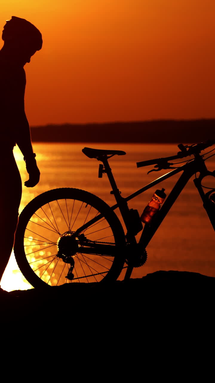 Modern bicycle on the background of the beautiful sunset. Bike near the river and the silhouette of a man wearing helmet and sport clothing in the evening. Vertical video
