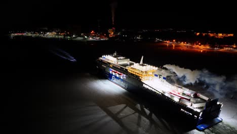 finnlines hybrid roro ship finnsirius moving through ice covered sea at night. searchlight illuminated. harbour lights ahead. moving aerial stern view.