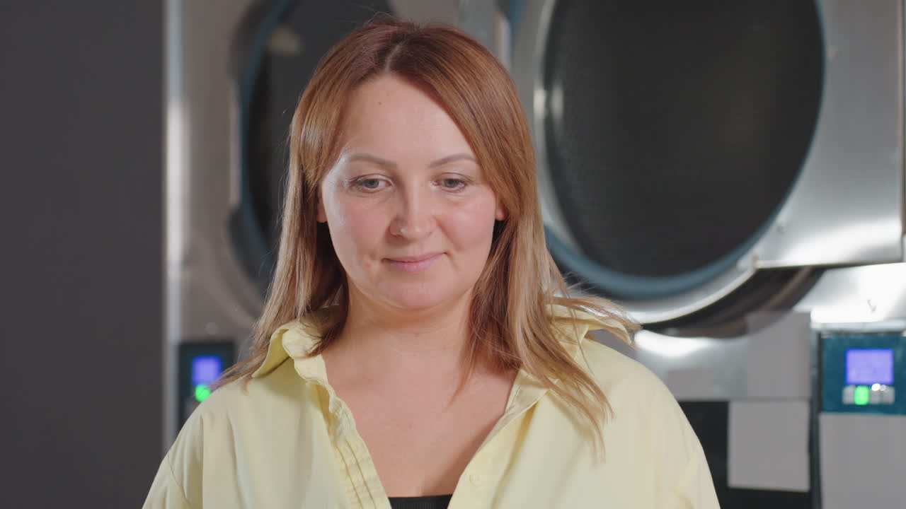 Portrait view of fair skinned laundry owner in yellow shirt, standing before stainless washers in modern laundromat, blur background, calm expression, professional business woman promoting hygiene