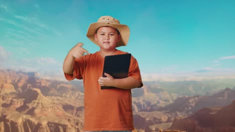 asian boy with a hat smiling and pointing to a tablet while traveling at the top of mountain. boy researcher examines something, travel tourism adventure concept
