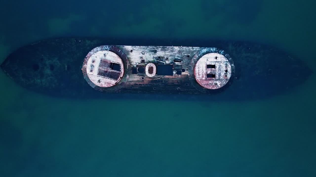 Aerial drone shot moving upward, revealing a dramatic shipwreck surrounded by ocean waves, captured from a bird's-eye perspective.