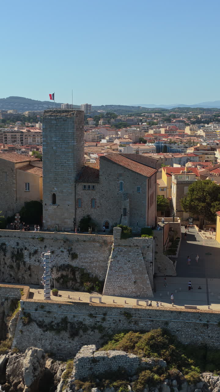 Aerial drone view of the Picasso Museum and medieval stone ramparts overlooking Antibes marina, with yachts docked and the Old Town's terracotta rooftops. Vertical