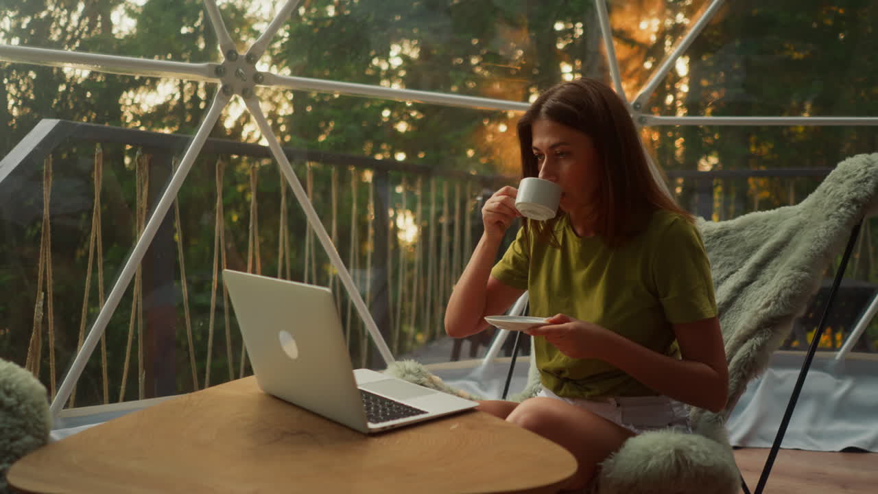 Woman distracts from work in online sphere for moment to drink cup of coffee. Young woman takes break holding plate with hand and looking at computer screen