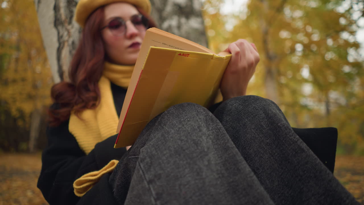 primer plano de un estudiante con gafas de sol, leyendo pensativo un libro cubierto de amarillo, las manos sujetan suavemente las páginas mientras el fondo presenta árboles de otoño borrosos