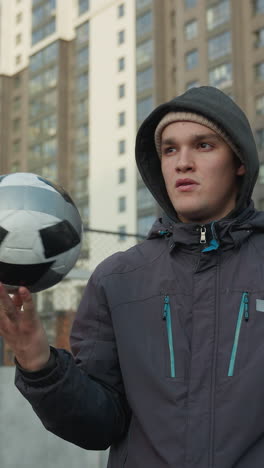 hombre con capucha de pie en un campo deportivo al aire libre gira la pelota de fútbol en su mano con enfoque y concentración, edificios urbanos de gran altura visibles en el fondo