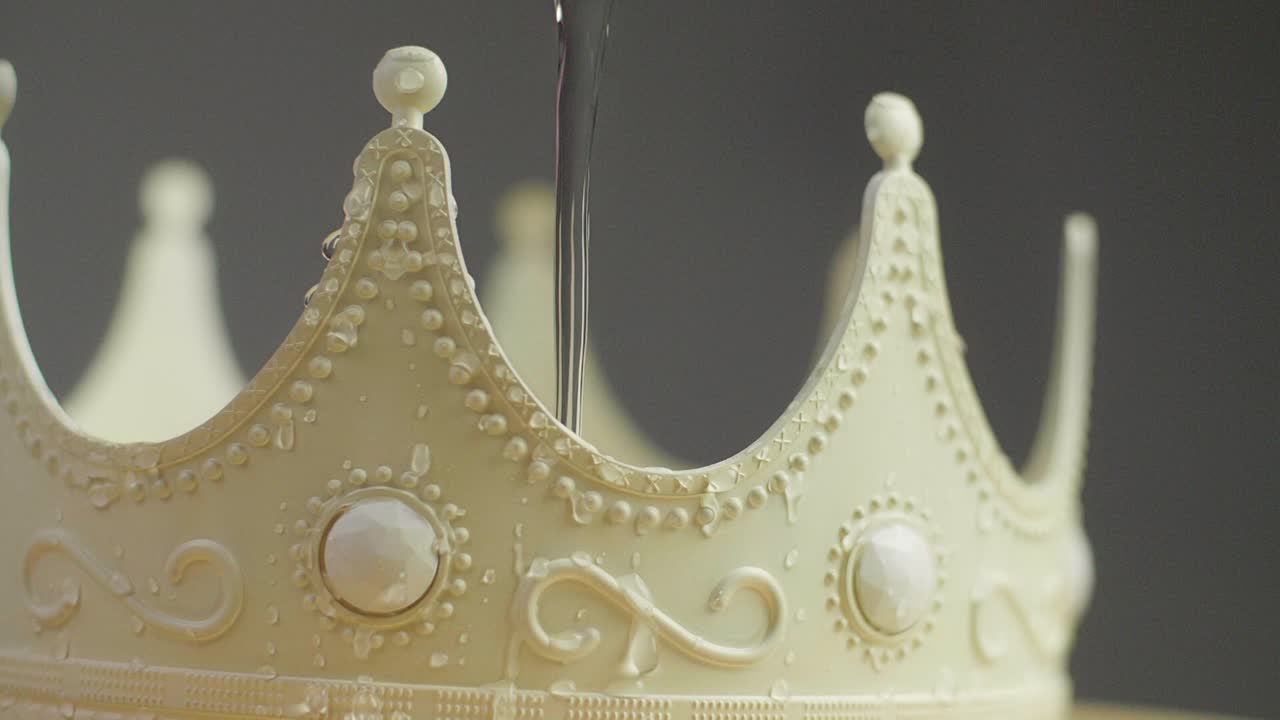 Close up of a beige crown on a pedestal with ornaments and white pearl jewels with peaks jagged out of plastic with water drops dripping on it. Smooth sliding camera movement. Shot in 4K.