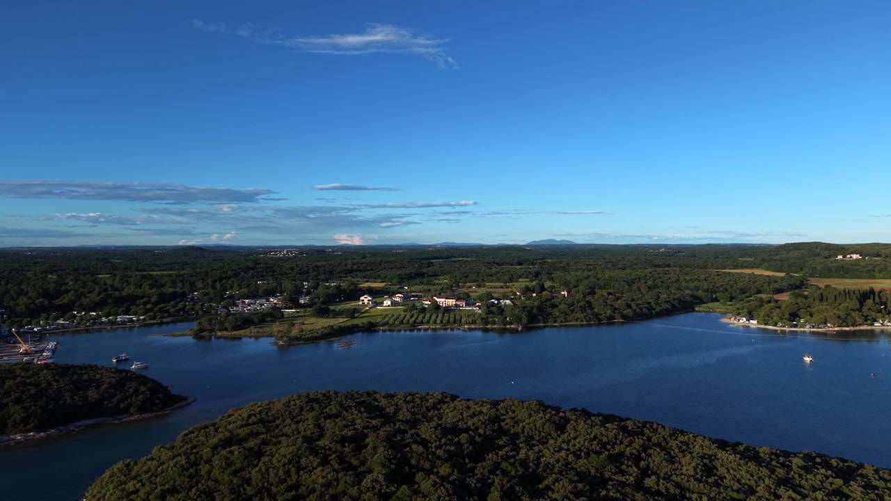 Forested island sits in calm blue channel near Istria, Croatia, with agricultural fields, vineyards and coastal village stretching across peninsula under clear summer sky. Aerial drone forward wide.