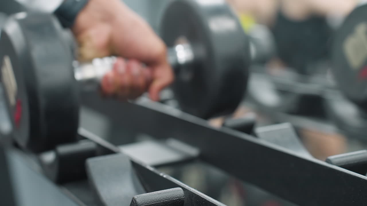 close of hand grabbing heavy black dumbbell from metal rack in well lit gym, showing preparation for strength training session with equipment neatly arranged in background