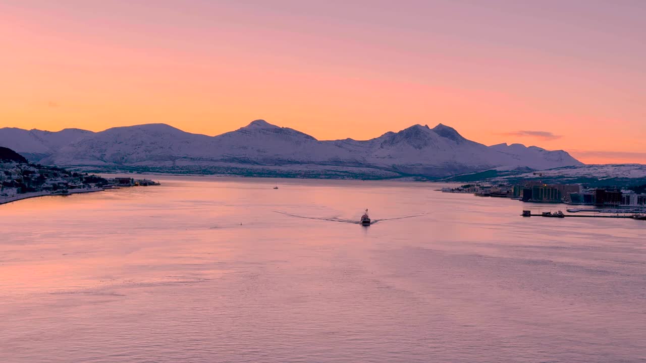 노르웨이의 트롬소 (tromso) 에서 눈 인 산 뒤에 해가 지고, 물 속에 있는 배가 배를 향해 오고 있다.