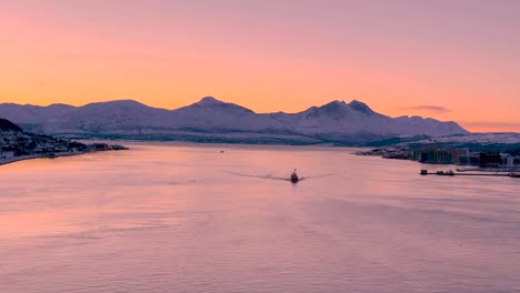 el sol se pone detrás de las montañas cubiertas de nieve en tromso noruega, con un barco en el agua que se acerca a los barcos