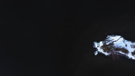 top down aerial footage flying sideways over a small snow covered island with the rocky bottom visible