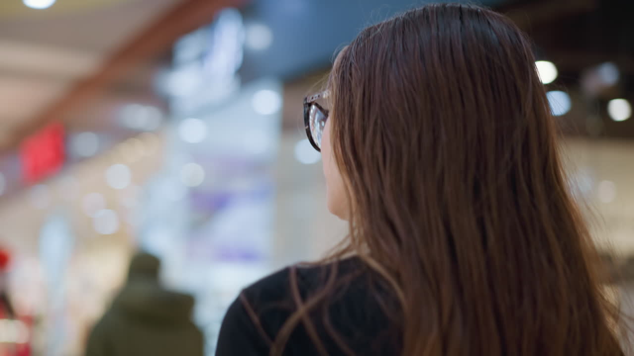 Rear view of lady in black top and glasses walking through mall, looking contemplative, background shows blurred person ahead with soft bokeh light effect