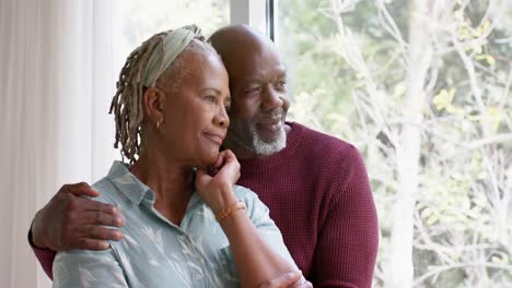 Reflexiva-Pareja-De-Ancianos-Afroamericanos-Abrazándose-Y-Mirando-Por-La-Ventana-En-Casa,-Cámara-Lenta