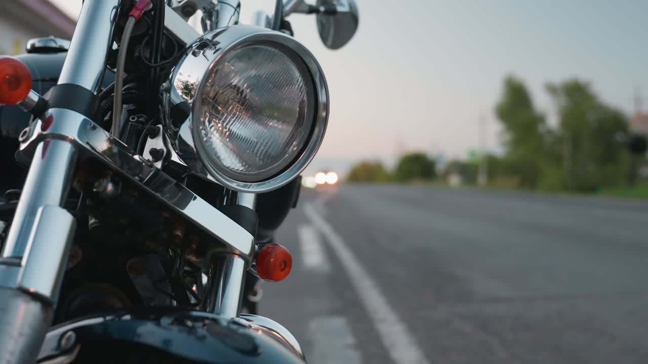 Parked motorcycle on roadside with chrome details reflecting evening light as passing car moves on lane and distant headlights glow softly against horizon creating travel mood