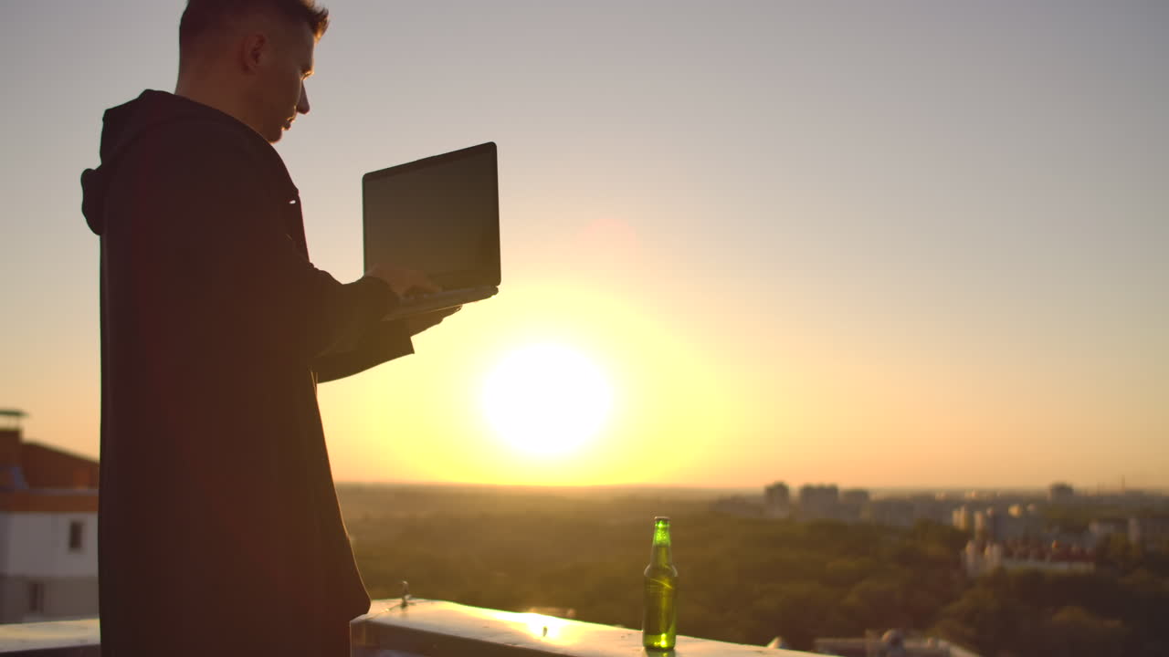 un programador con una computadora portátil se sienta en el techo y escribe código al atardecer. trabajo remoto freelance. libertad para trabajar. escribir en un teclado al atarrear el sol con una vista de la ciudad.