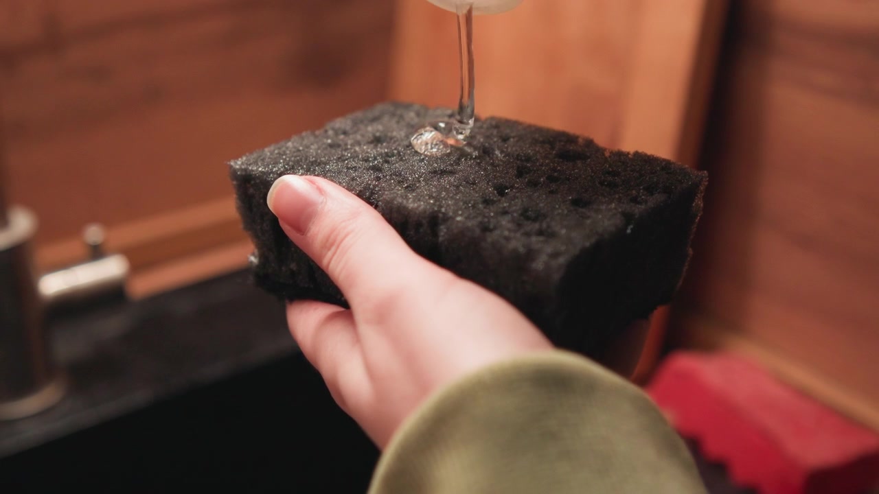 La mano destra tiene una spugna scura testurizzata sopra il lavello, strizzando l'acqua mentre schiuma di sapone e goccioline si raccolgono nelle perforazioni; un visibile movimento di gocciolamento e le punte delle dita lucide suggeriscono una pulizia di routine.