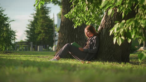 lady sitting under tree in grassy field, reading book with sunlight casting a warm glow, background features soft blur of light poles and building, surrounded by greenery