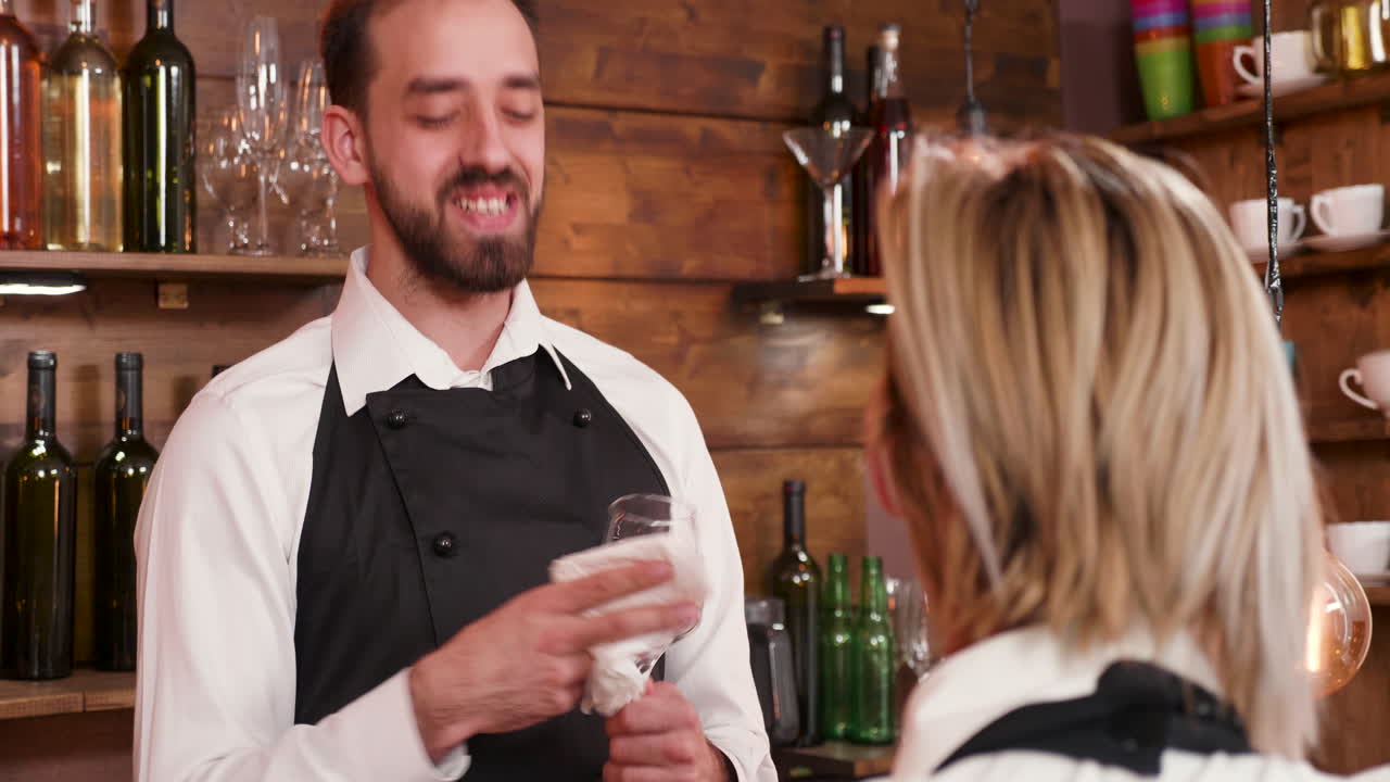 A bartender wipes a wine glass behind the counter while serving a customer