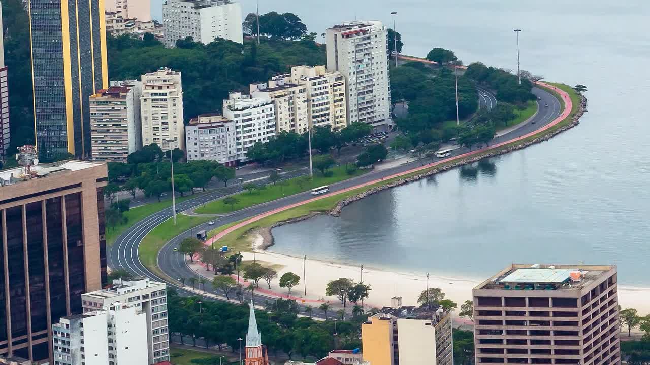 Filmato in time-lapse di una scena urbana sul lungomare che presenta una strada curva che costeggia una baia. La strada è delimitata da spazi verdi, alberi e una zona di spiaggia sabbiosa, mentre grattacieli