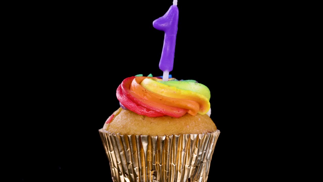 Hand inserts purple number one candle into rainbow-frosted cupcake with gold wrapper, black background