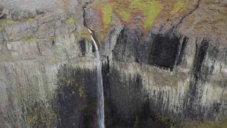 Toma-Aérea-Hacia-Atrás-De-Una-Cascada-Que-Cae-En-Un-Paisaje-Volcánico-De-Islandia-Durante-El-Día.