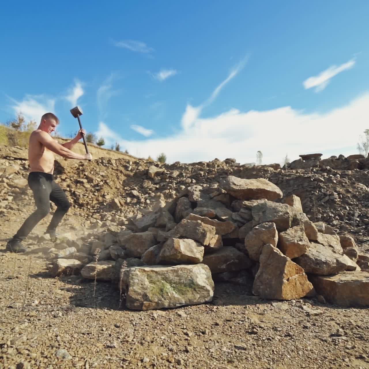 Sportsman with hard hammer on the rocky background. Athletic man without shirt breaking stones with metal hammer outdoors. Slow motion.