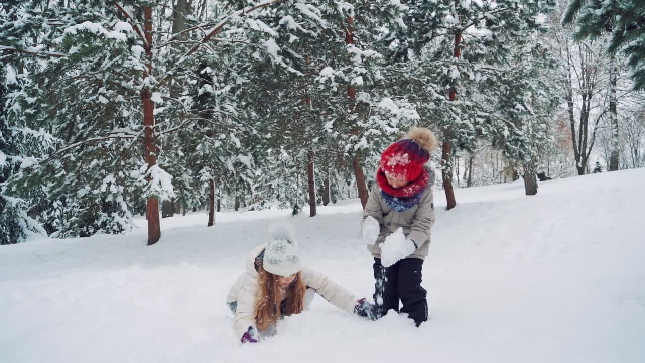 Funny children in warm clothes and hats are forming a large snowballs in their hands in the park in the winter on the background of green fir-trees. Slow motion