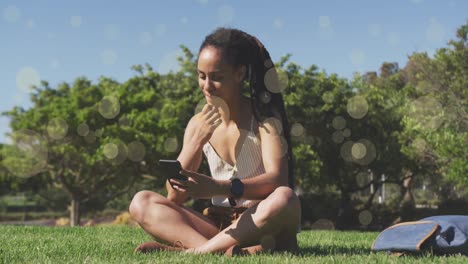 manchas amarillas cayendo contra una mujer afroamericana usando un teléfono inteligente sentada en la hierba