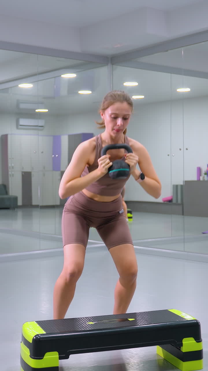 mujer levanta pesas, camina en un banco de aeróbico, luego baja y levanta peso de nuevo en un gimnasio con paredes de espejo, entrenamiento de fuerza y rutina de ejercicio cardio que combina la construcción muscular