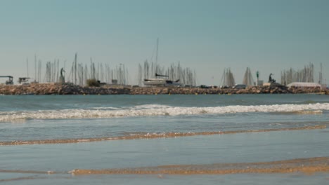 Toma-En-Cámara-Lenta-De-Olas-Rompiendo-En-La-Playa-De-Palavas-Con-Un-Puerto-Deportivo-Detrás