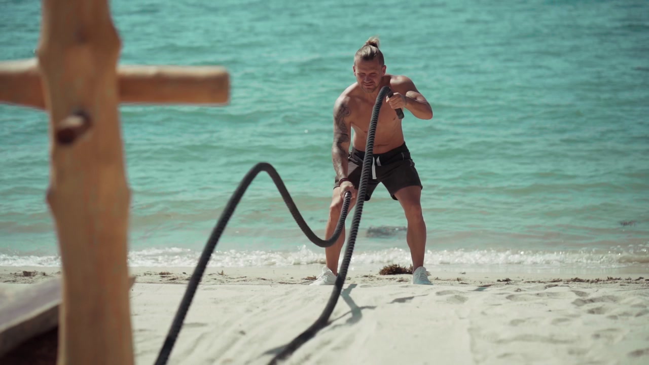 Uomo muscoloso che si allena con le corde da battaglia su una spiaggia soleggiata