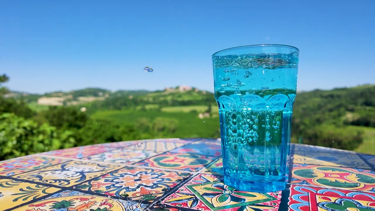 Carbonated water fills a blue glass on a mosaic table against a sunny countryside landscape.