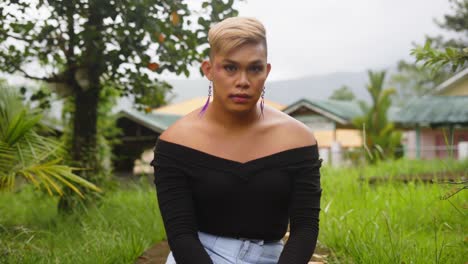 gay asian man with short blonde hair and boho earrings, wearing black off-shoulder top and sitting outdoor