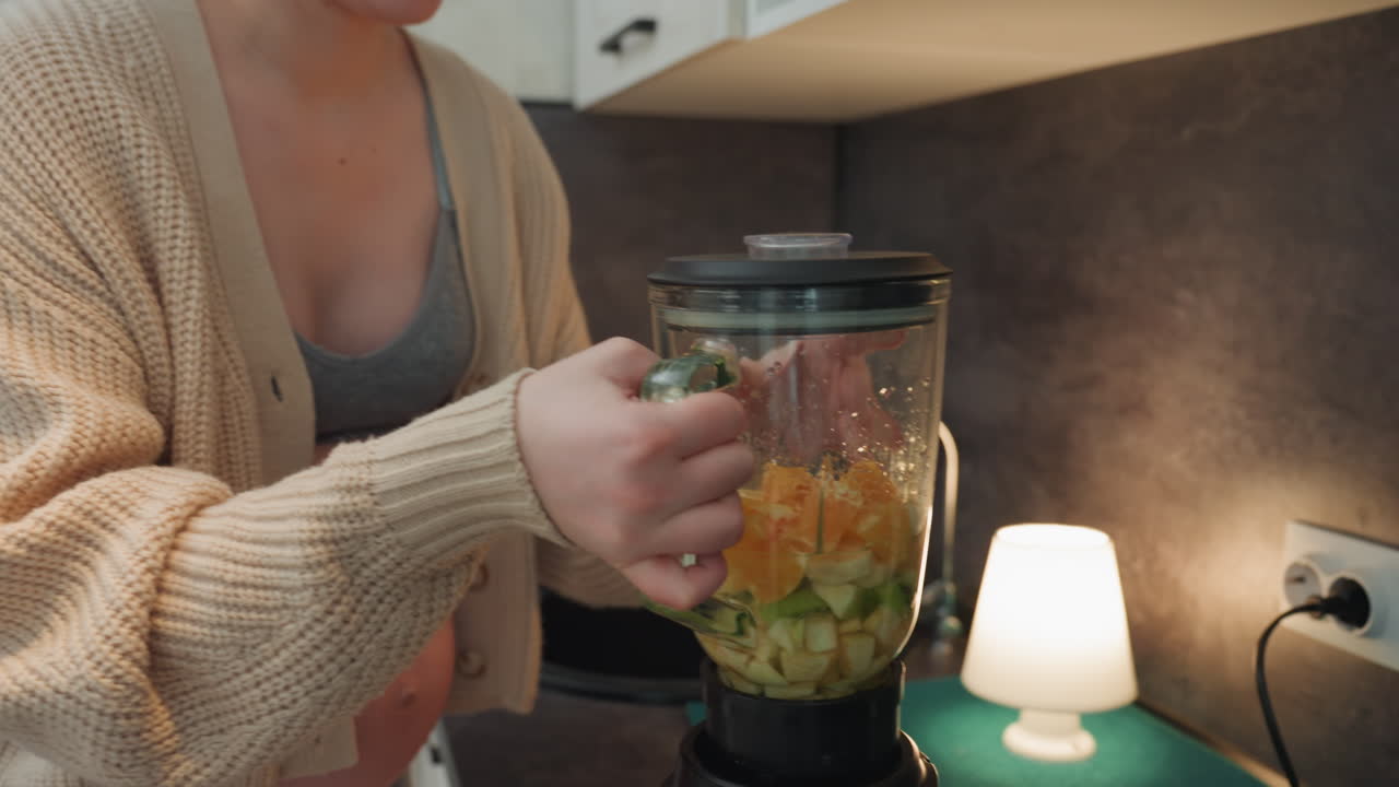 Madre caucasica incinta che fissa il coperchio del frullatore, mani ferme che posizionano il tappo sul barattolo, pancia scoperta e cardigan, bagliore della lampada sul piano di lavoro, ingredienti ricchi di vitamine pronti, rituale misurato dello spuntino prenatale
