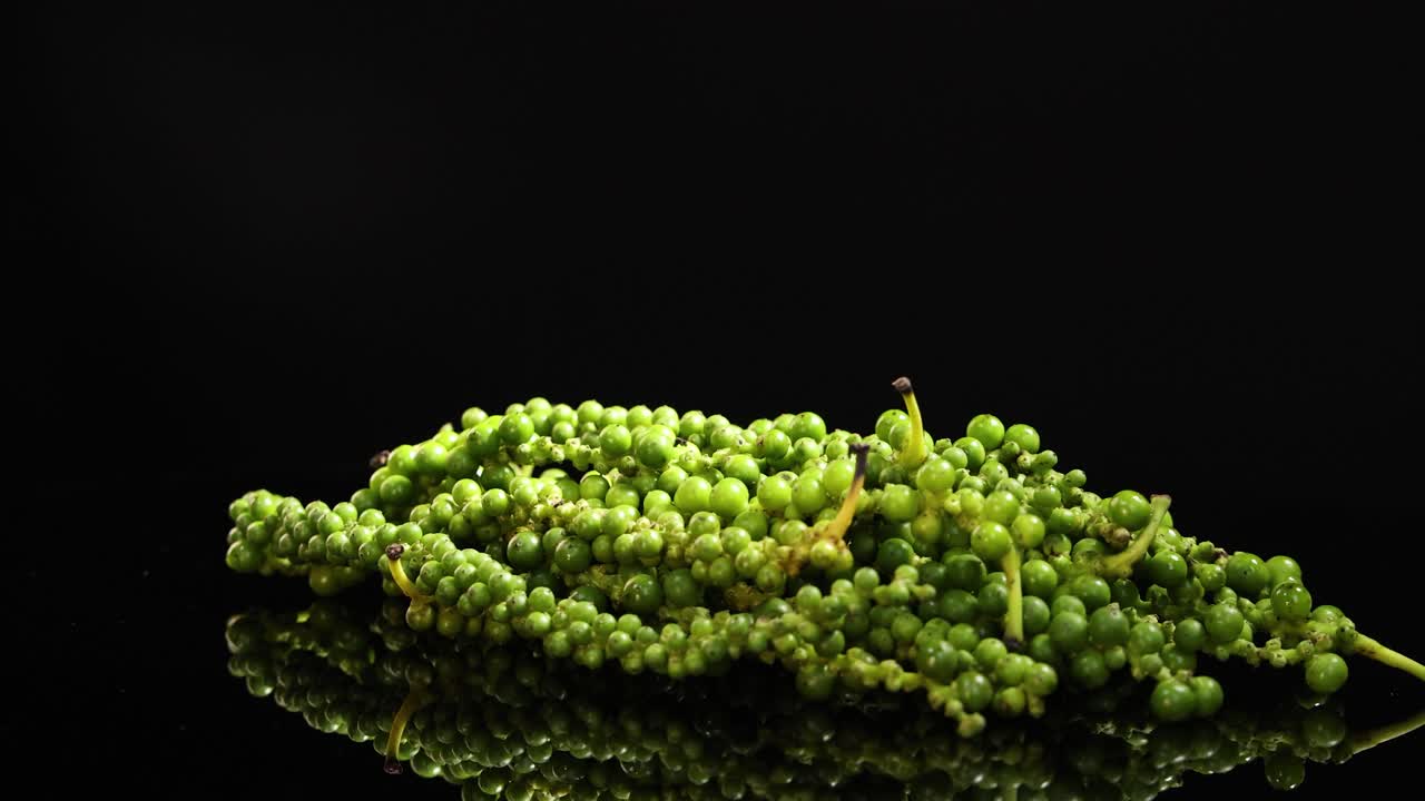 A cluster of fresh green peppercorns rotates smoothly on a reflective black surface, illuminated by dramatic studio lighting for a vivid, high-contrast effect