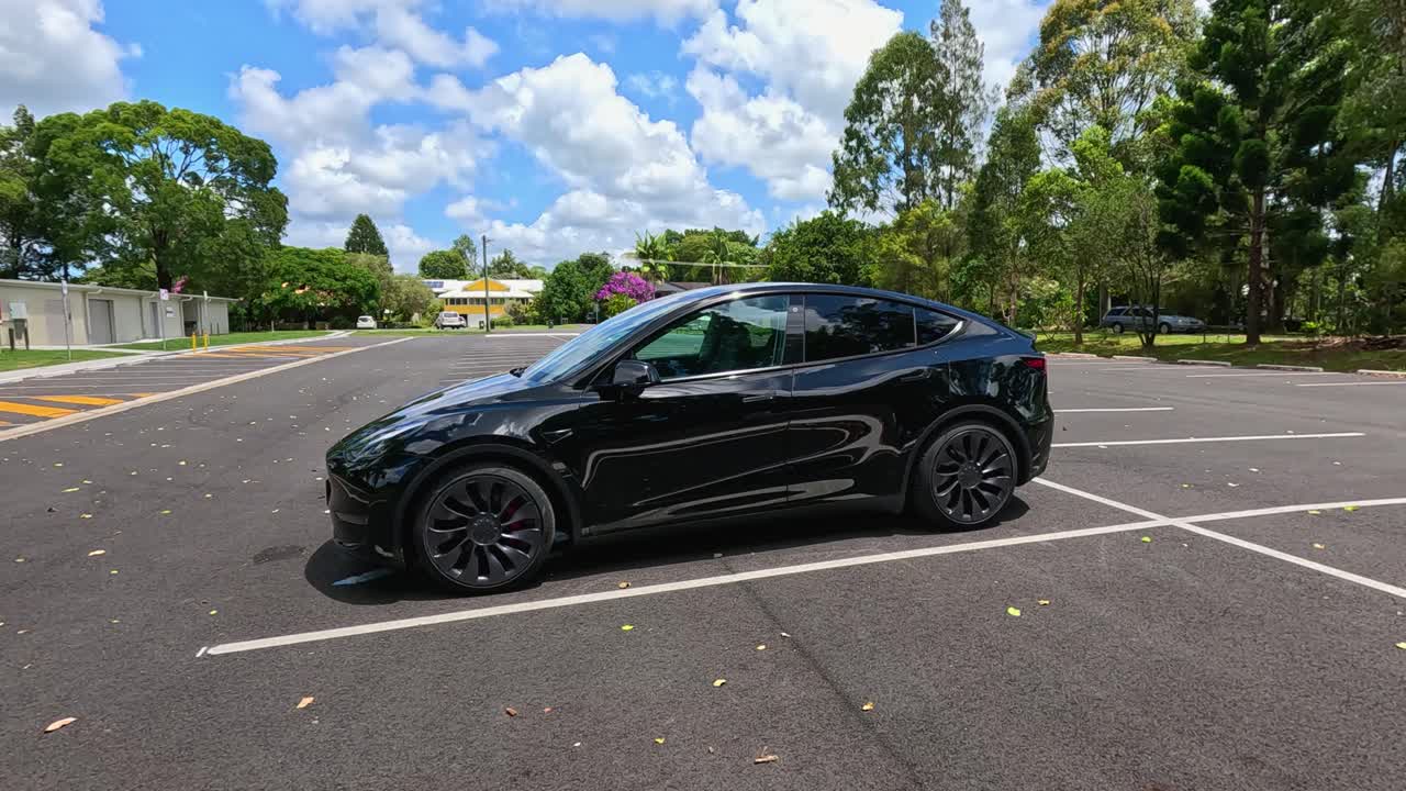 Black Tesla Model Y parked in an empty lot under bright daylight, surrounded by trees and clear skies