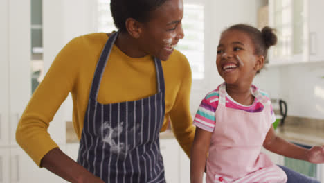 Happy-african-american-mother-and-daughter-wearing-aprons-having-fun-while-cooking-in-kitchen