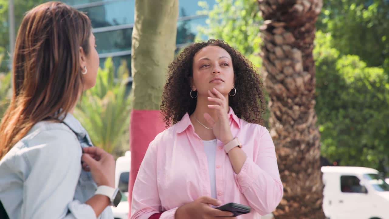 Afroameričanky stojí na ulici, telefon bzučí, pravá žena prohlíží obrazovku a levá natahuje ruku.